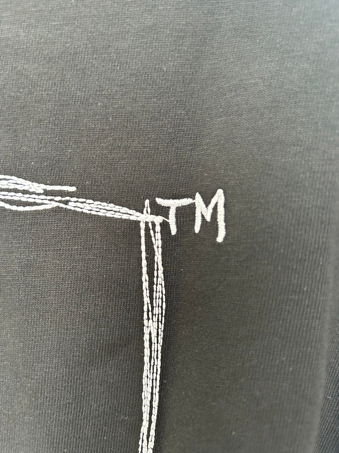Black T-shirt back detail with white embroidered-style “TM” text