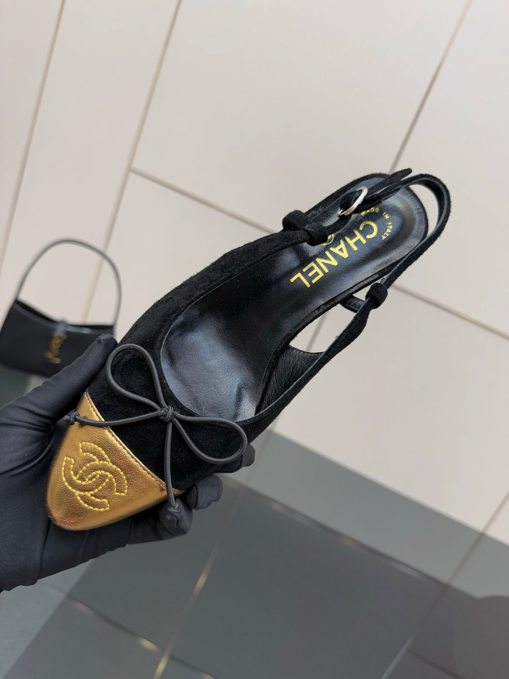 Hand holding black suede ballet flats with gold toe cap and bow laces, side view.
