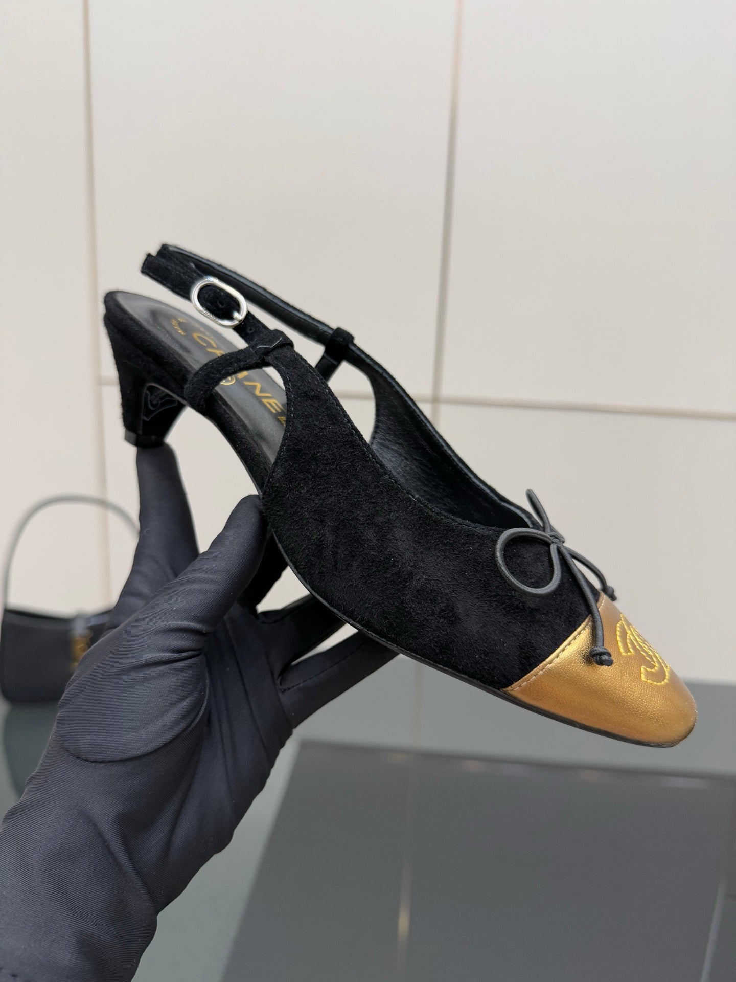 Hand holding a black suede ballet flat with gold toe cap and bow lace.