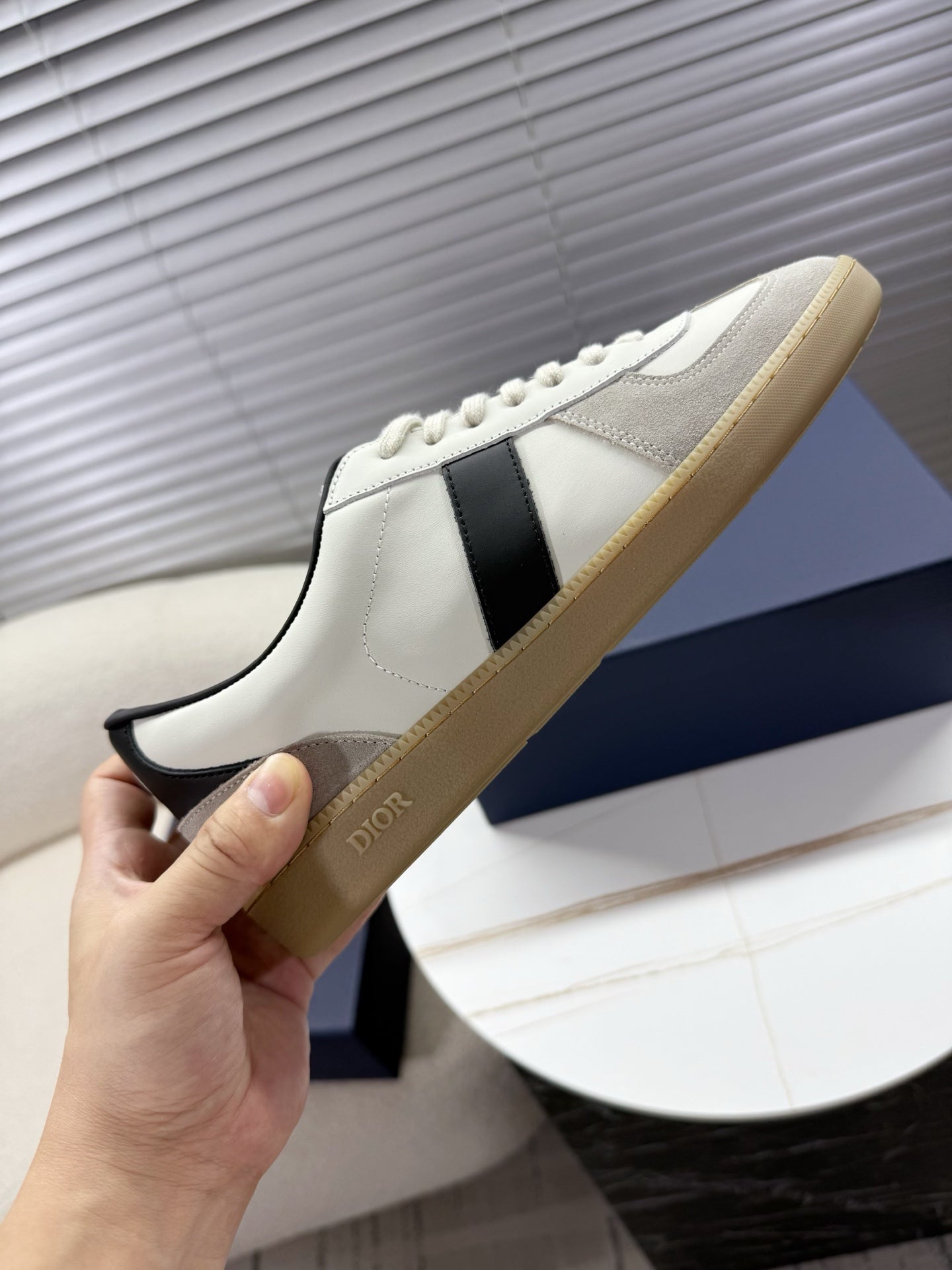 Hand holding white sneakers with black side panel, cream sole, lace-up front