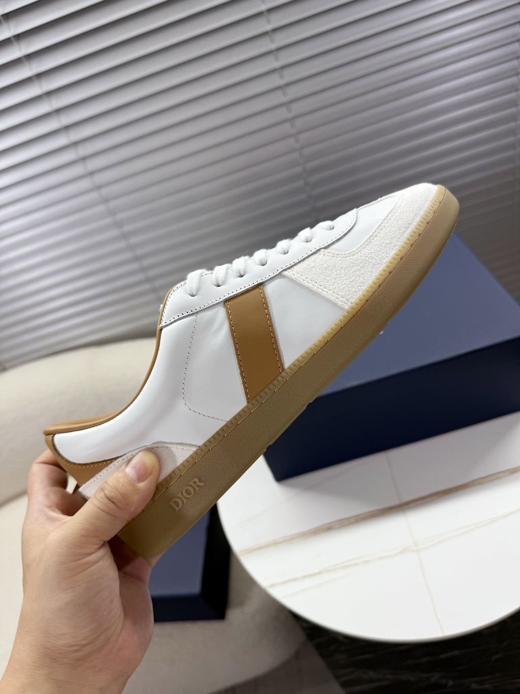 White Dior sneaker held by hand; tan side stripe and gum-style sole