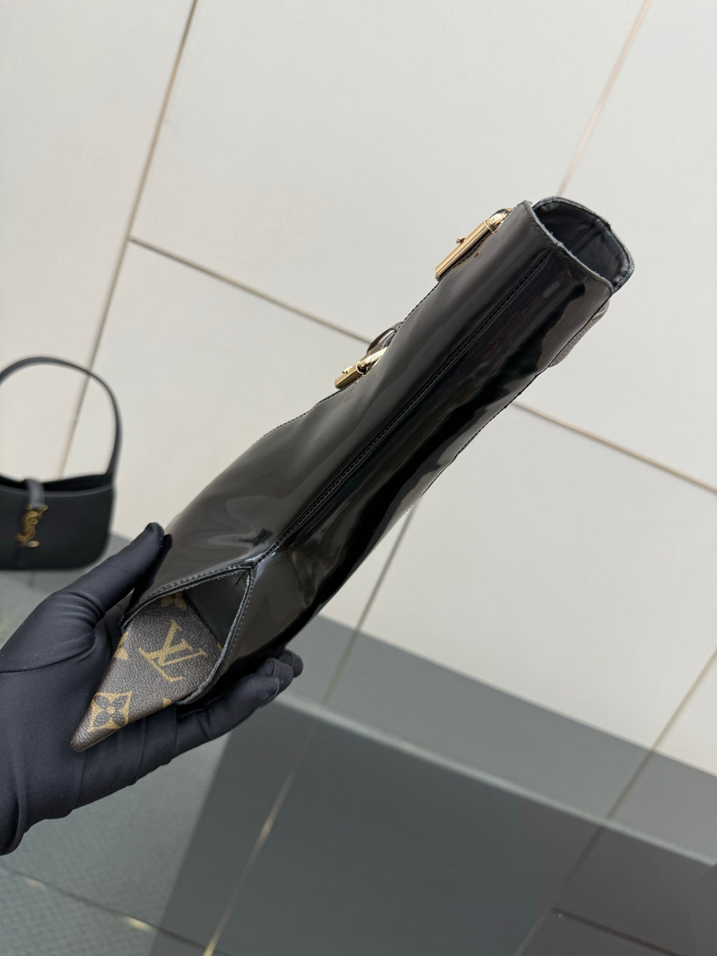 Black patent ankle boot held in hand, side profile with gold hardware.