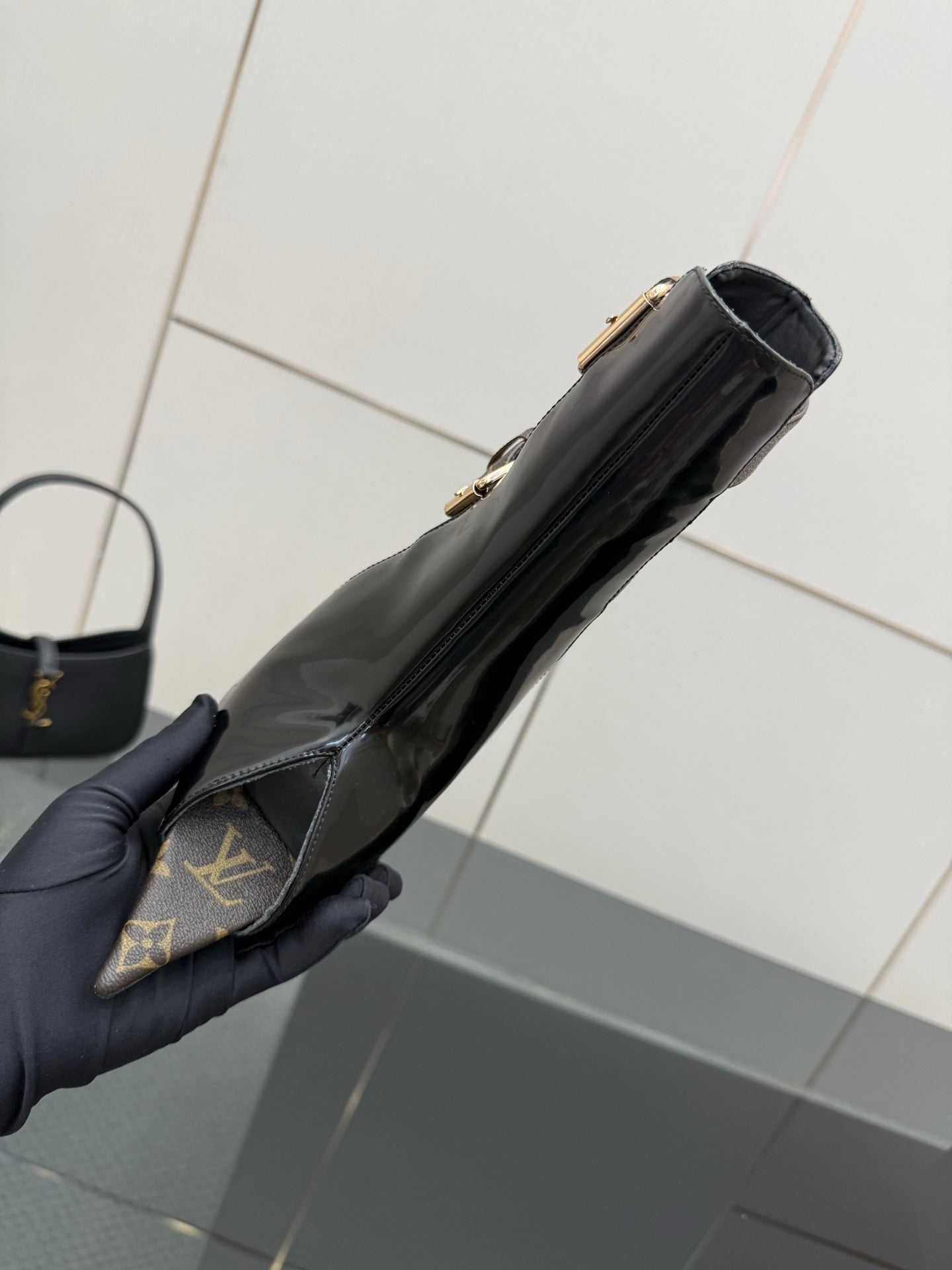 Black patent ankle boot held in hand, side profile with gold hardware.