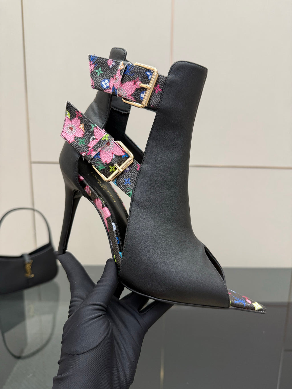 Close-up black ankle boot with floral strap, gold buckle, and pointed toe heel.
