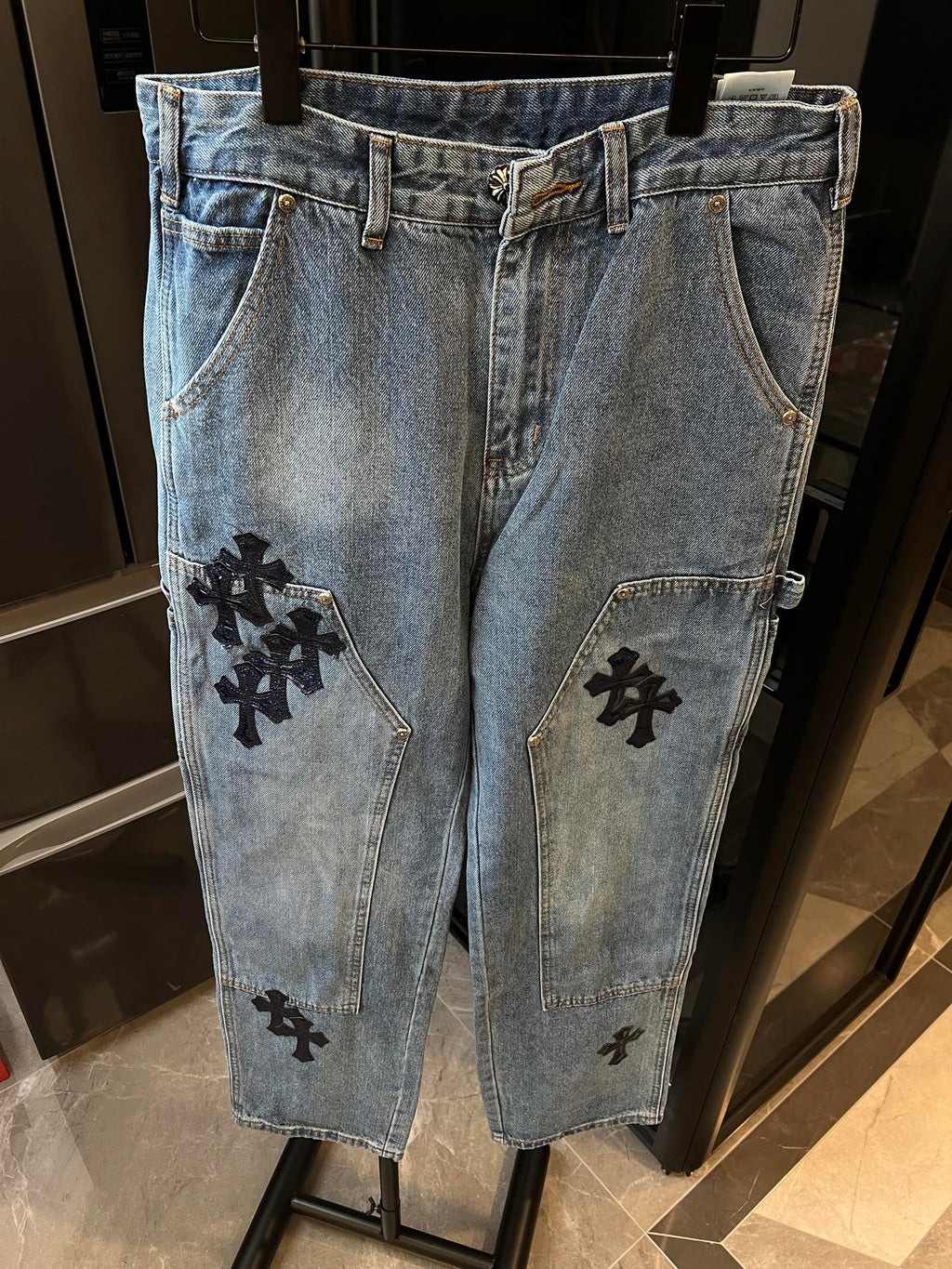 Light-wash denim pants with black cross patches on front pockets and leg panels