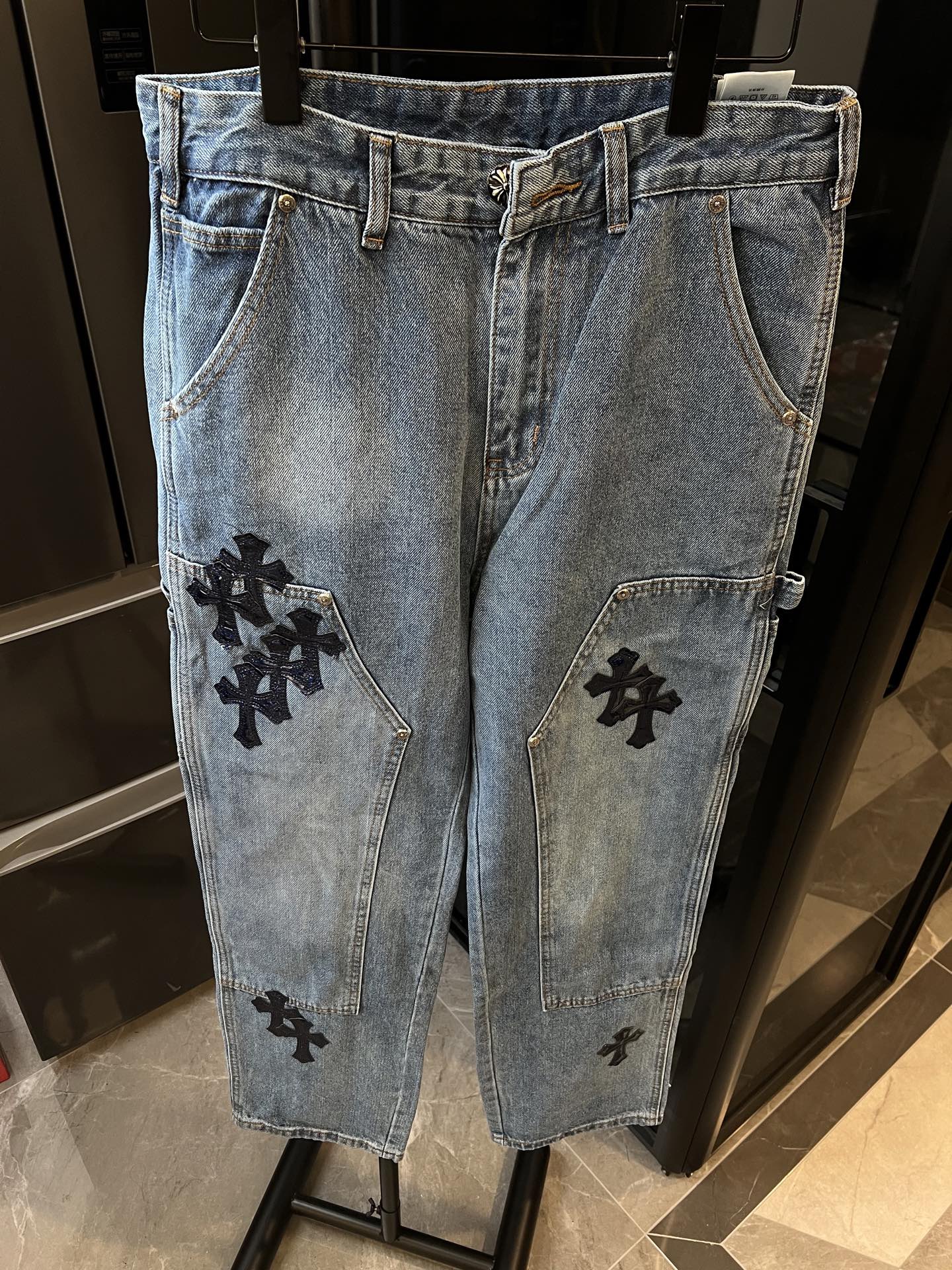 Light-wash denim pants with black cross patches on front pockets and leg panels