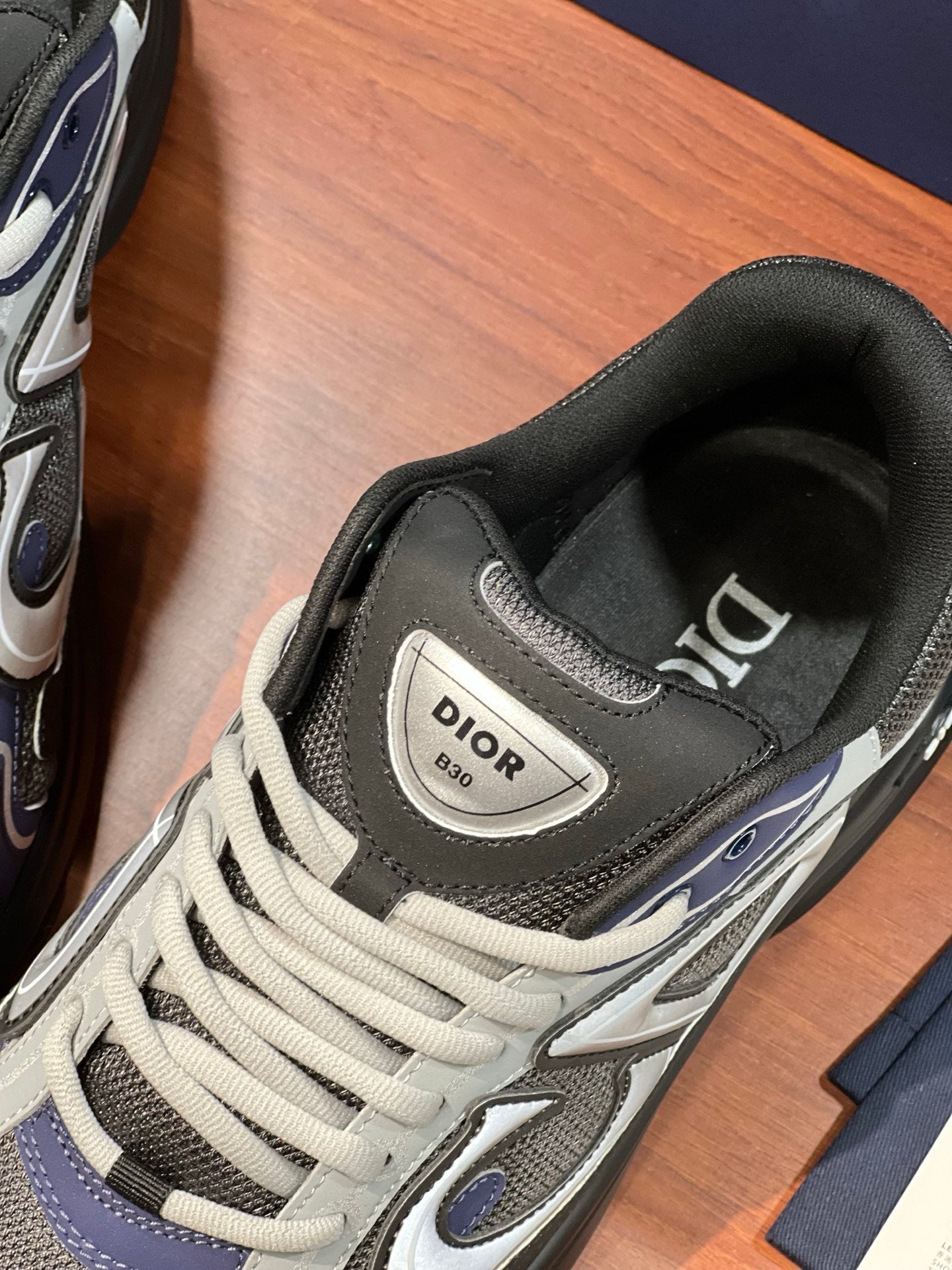Sneaker close-up: Dior logo heel patch, gray upper, white laces, navy trim.