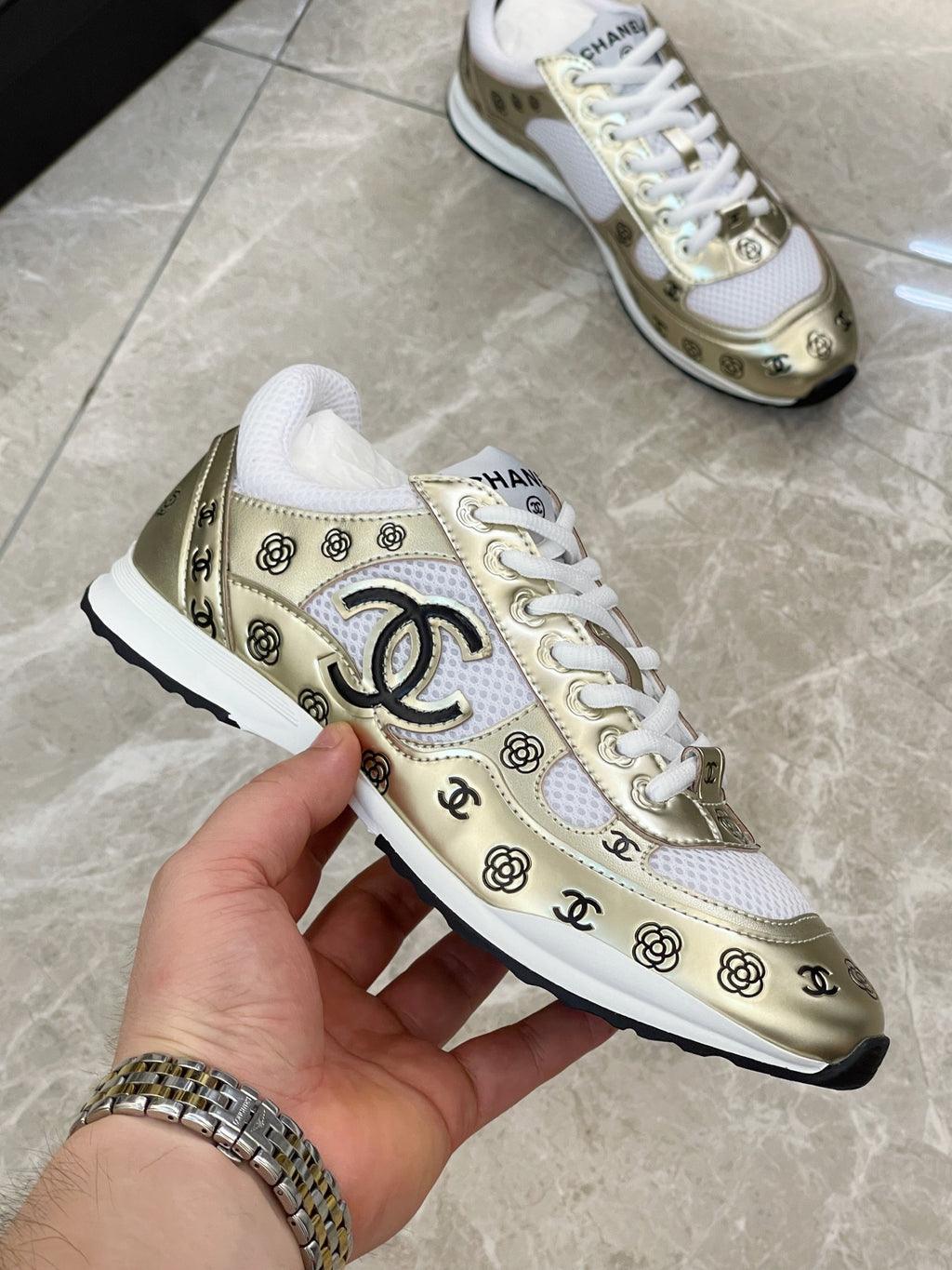 Unisex sneakers held in hand, gold and white logo pattern, white laces