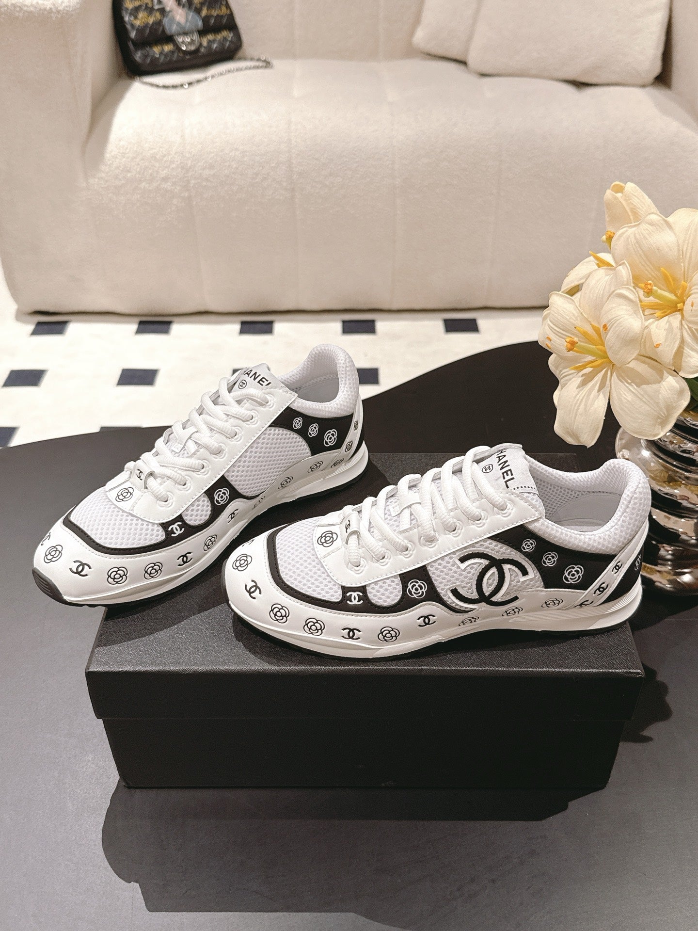 White low-top sneakers with black trim and repeating Chanel CC logos, pair on box