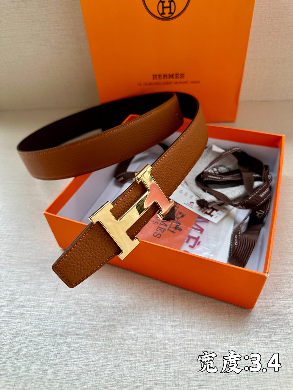 Leather belt with brown strap and silver-tone H buckle, in orange gift box setup.