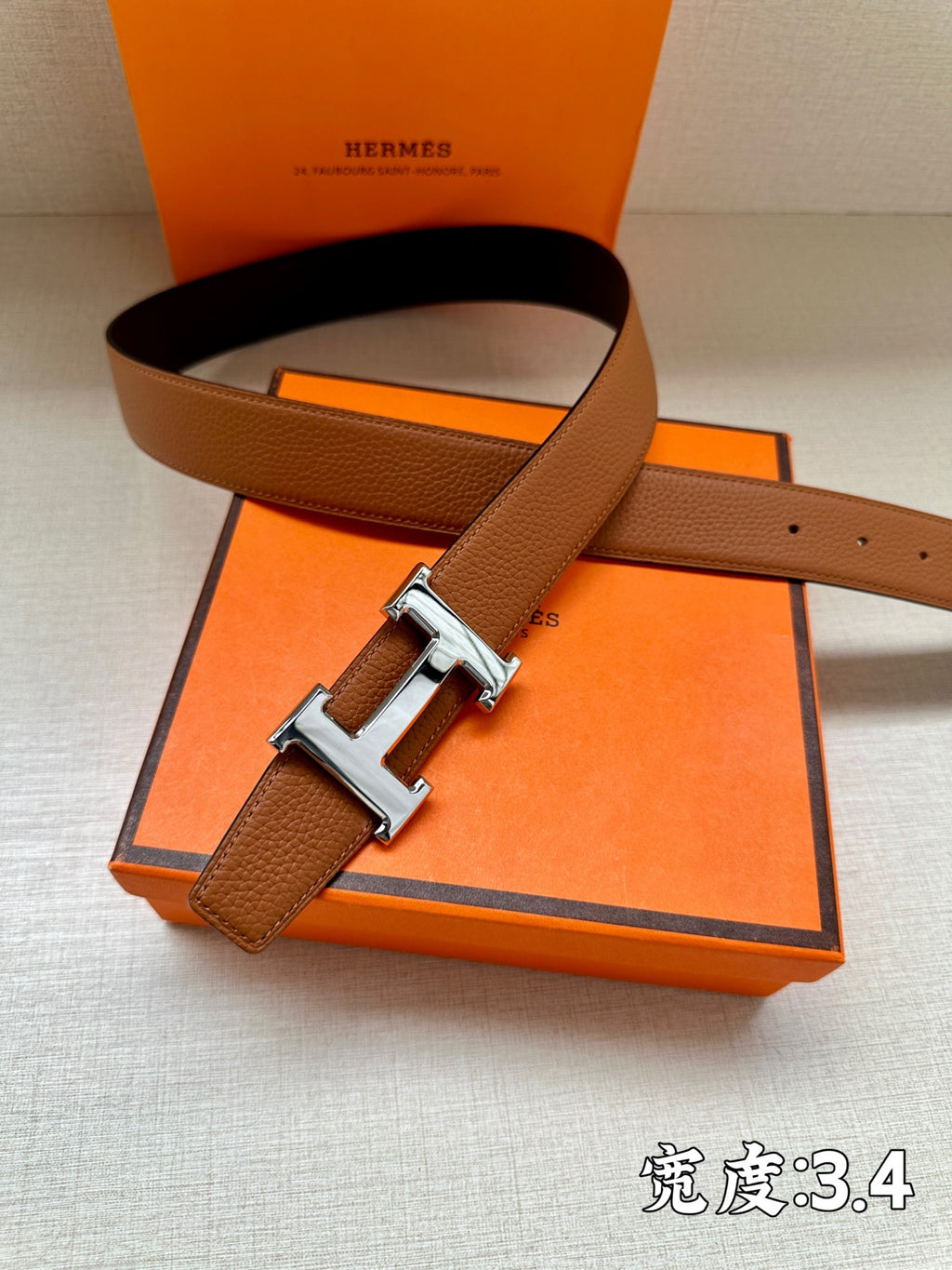 Leather belt with brown strap and silver-tone H buckle, shown over orange gift box.