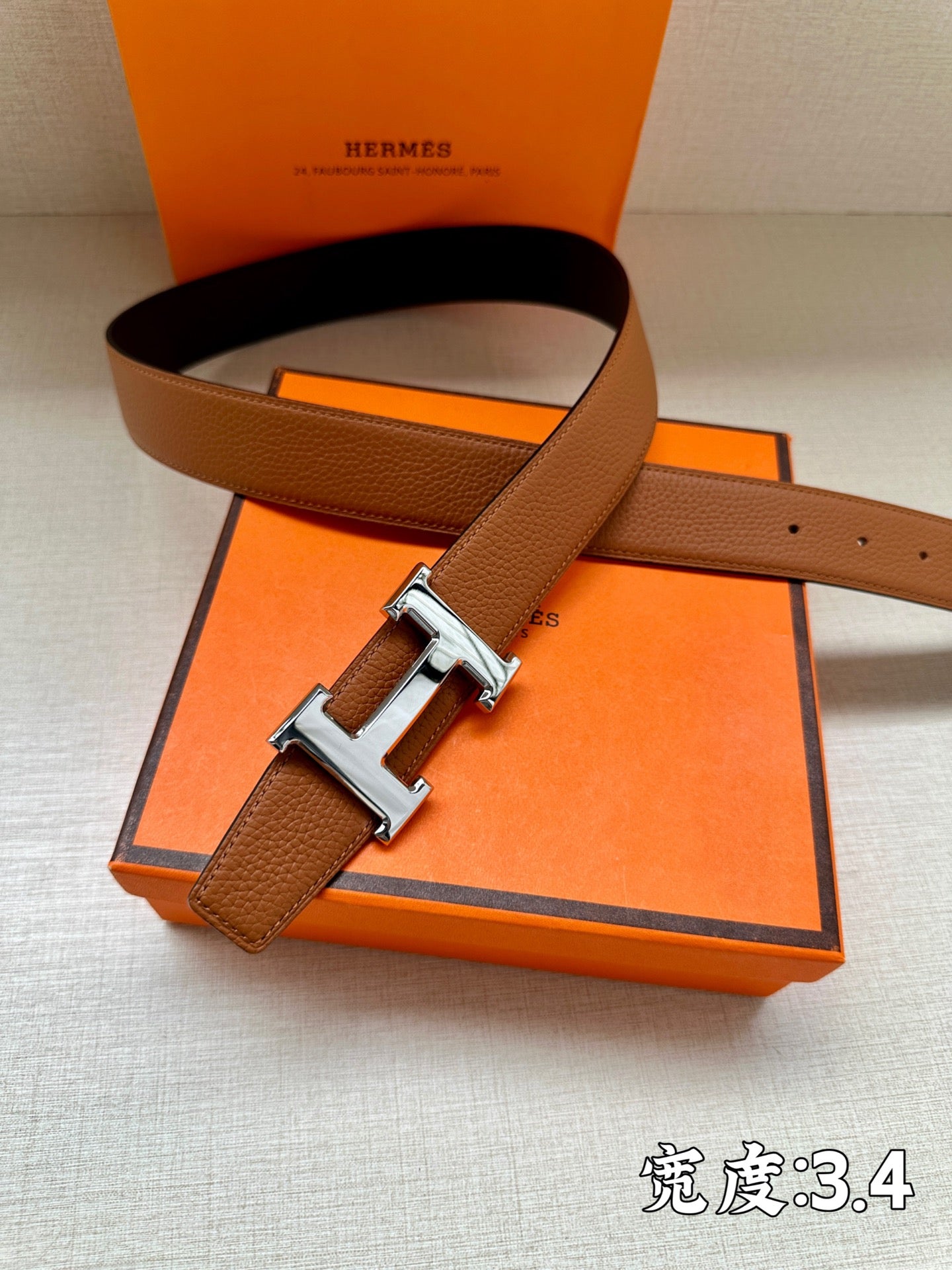 Leather belt with brown strap and silver-tone H buckle, shown over orange gift box.