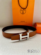 Leather belt with brown strap and silver-tone H buckle, laid flat with orange packaging.