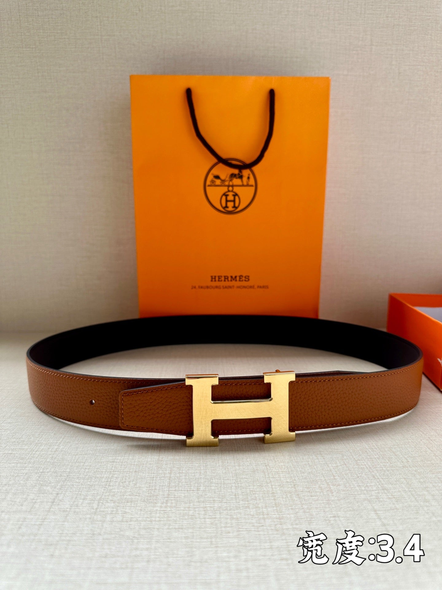 Leather belt with brown strap and silver-tone H buckle, laid on fabric with box behind.