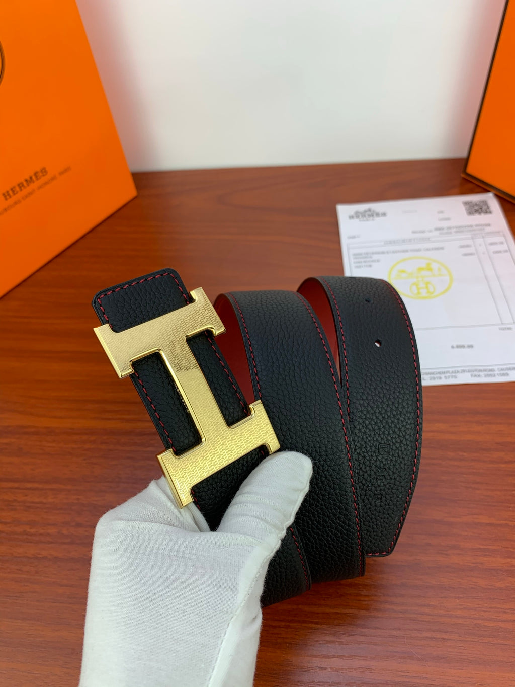 Black leather belt with gold H buckle and red edge trim, held in a glove.