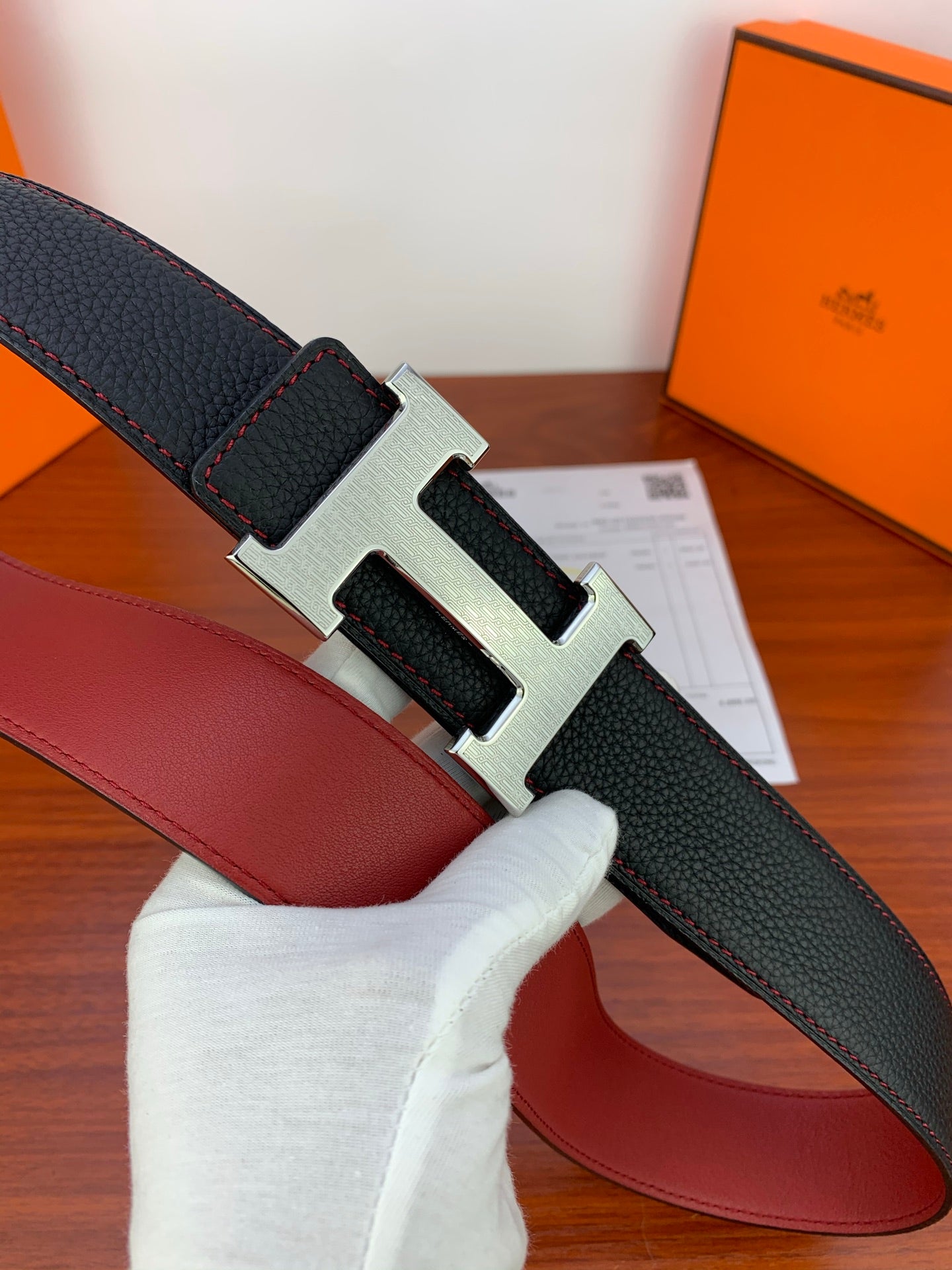 Black leather belt with silver-toned H buckle and visible red underside, close-up.