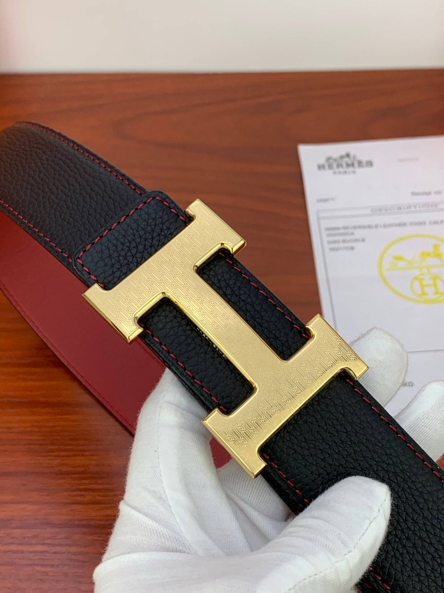 Black leather belt with gold H buckle detail and red contrast stitching, close view.
