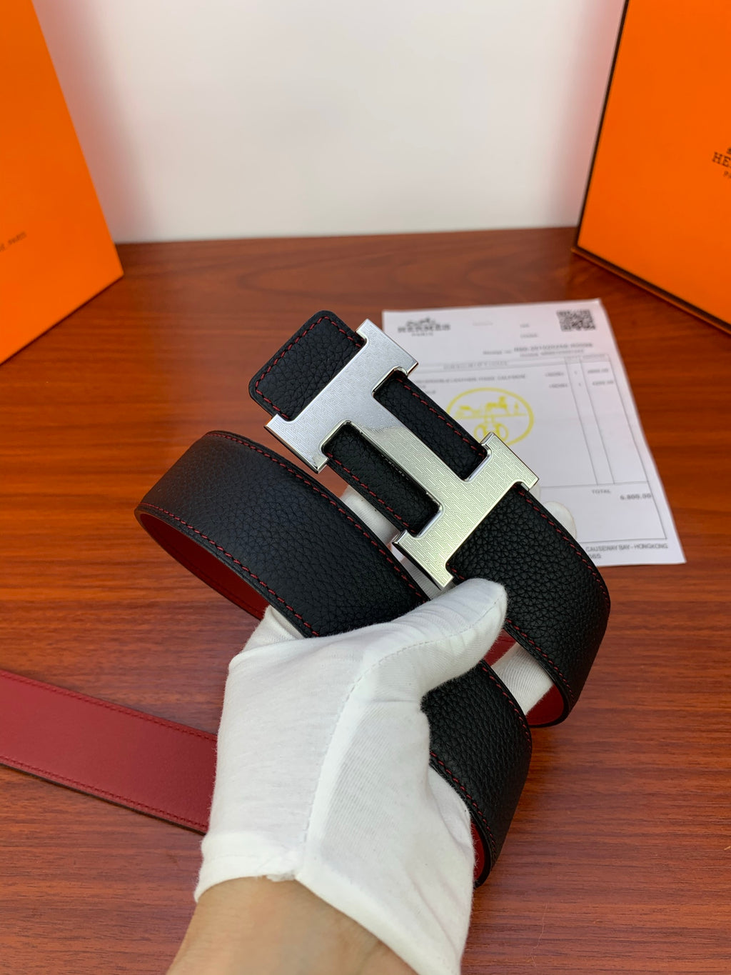 Black leather belt with gold H buckle and red interior panel, angled view.