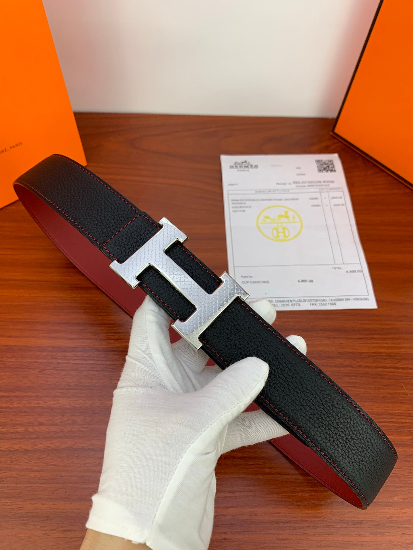 Leather belt with black textured strap and silver-tone H buckle, held in a gloved hand