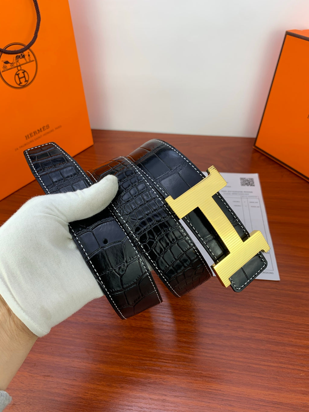 Dark croc-embossed leather belt with gold-tone buckle held in a glove.