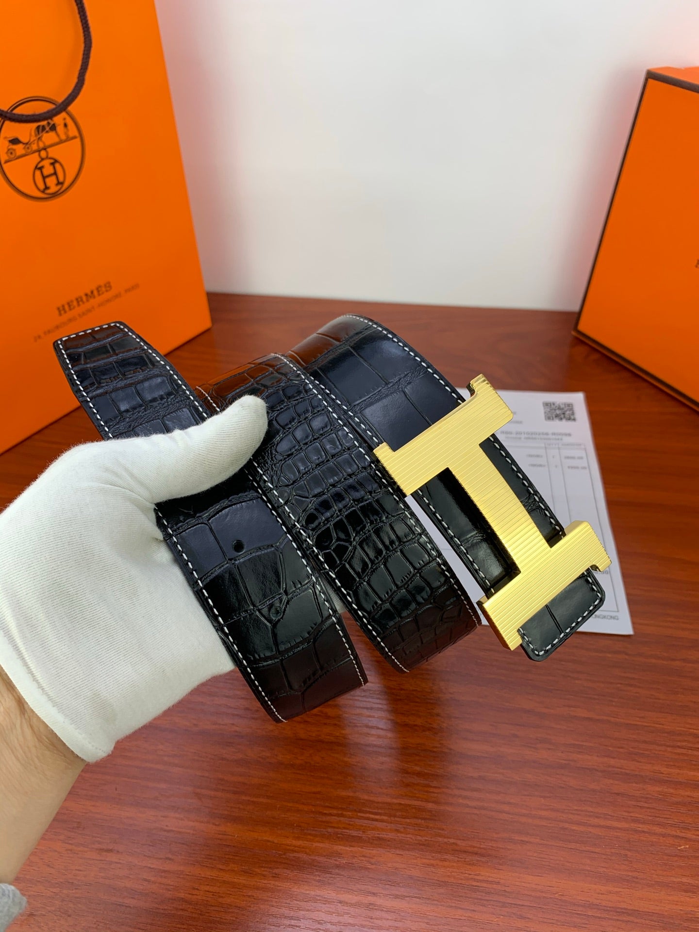 Dark croc-embossed leather belt with gold-tone buckle held in a glove.