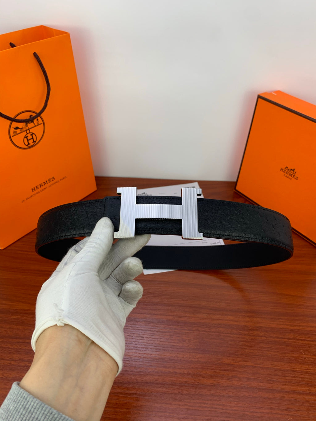 Black leather belt with gold H buckle, laid flat and shown in gloved hand