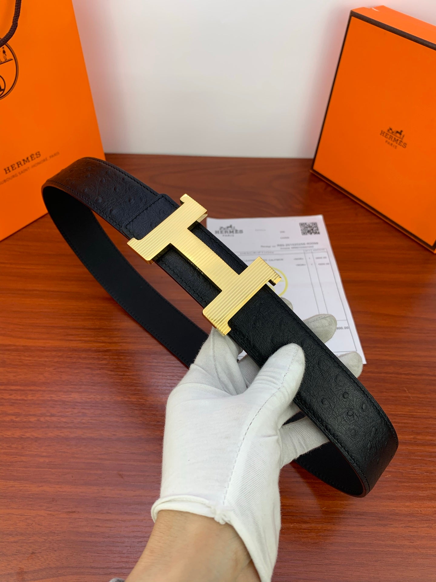 Black leather belt with gold H buckle, held in gloved hand