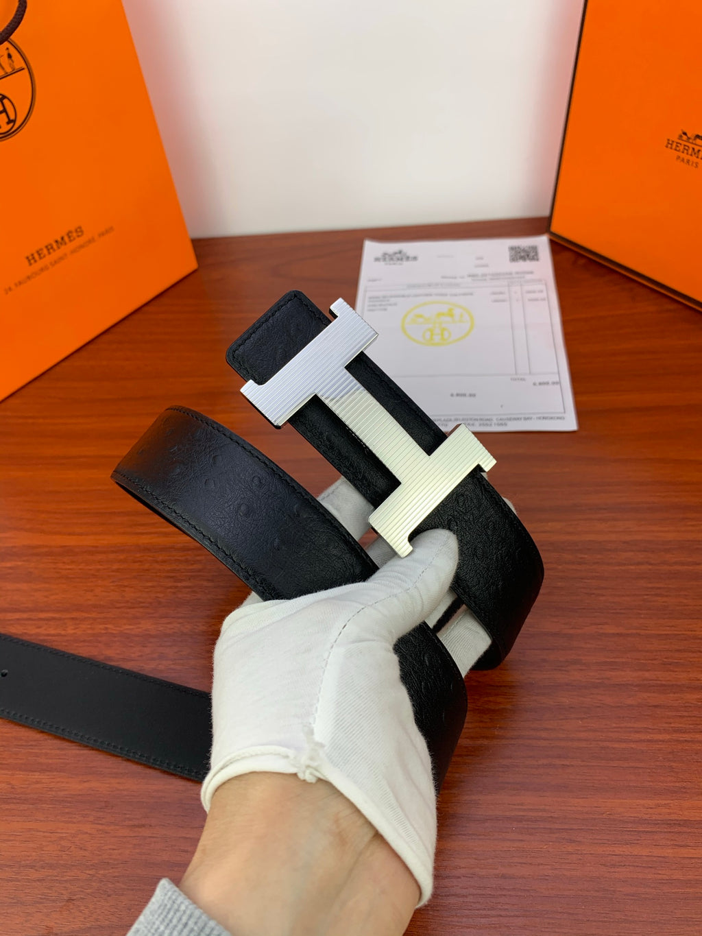 Black leather belt with gold H buckle, photographed at an angle in gloved hand