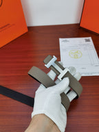 Hermes belt with light gray leather strap and gold-tone H buckle held by glove
