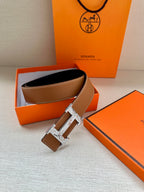 Tan-brown leather belt with silver H buckle, laid on orange box