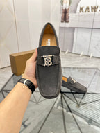 Dark gray suede loafer held up by hand, featuring B logo strap and tan lining