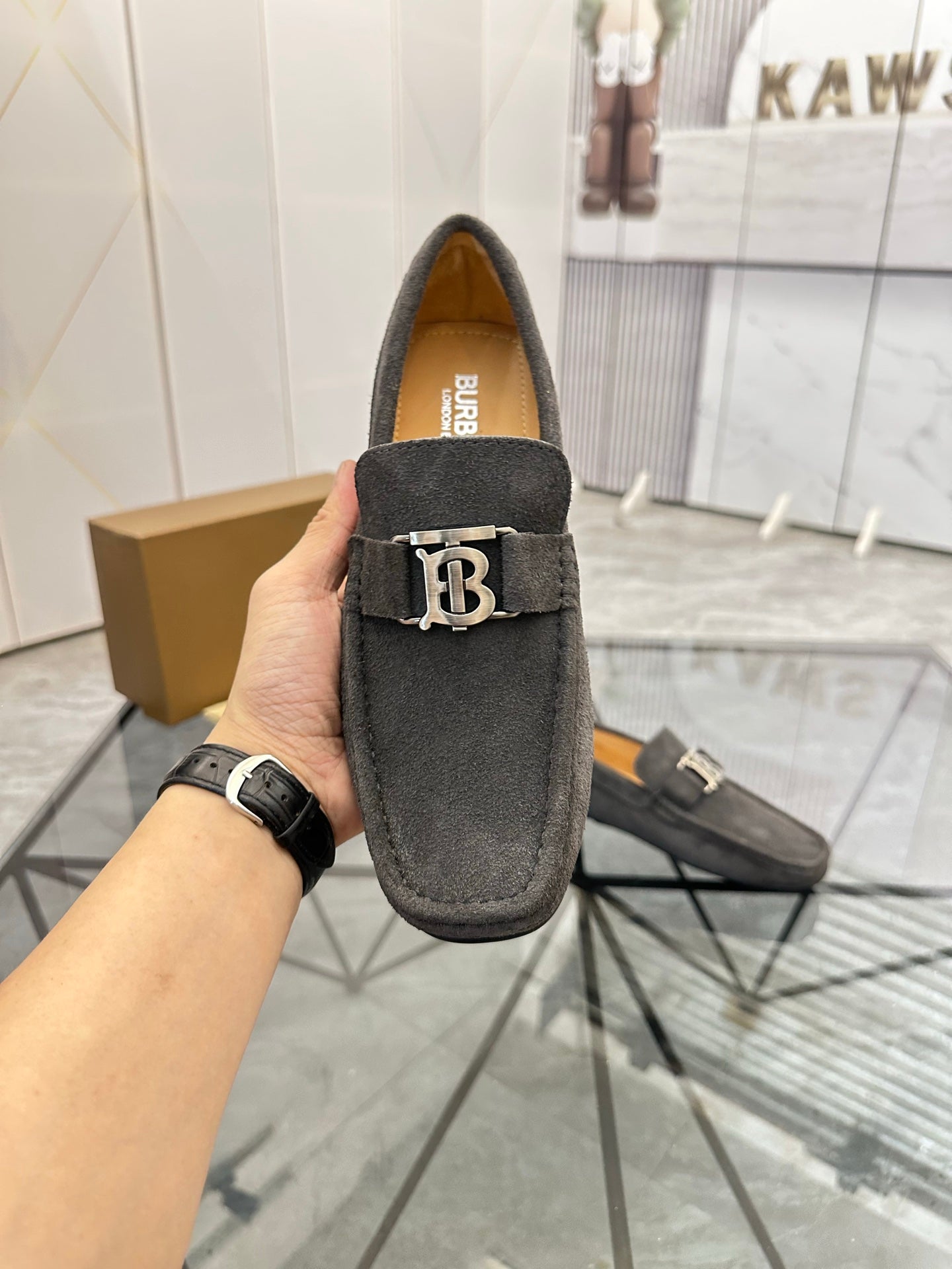 Dark gray suede loafer held up by hand, featuring B logo strap and tan lining