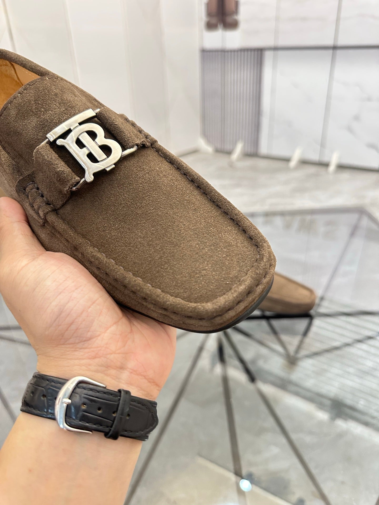 Brown suede loafer close-up with white B buckle and stitched moccasin-style edge.