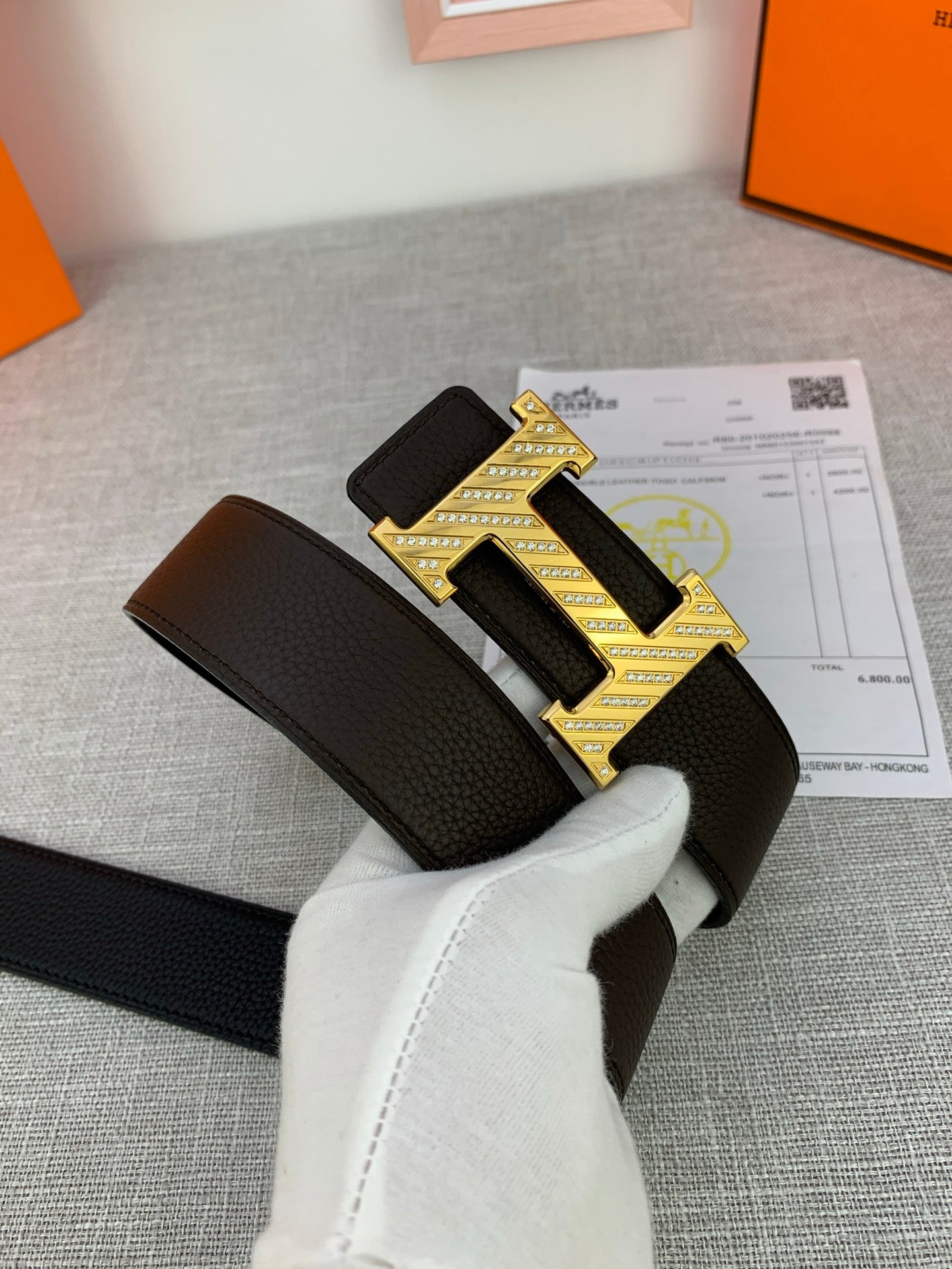 Hermes belt with dark leather strap and gold patterned buckle, held by white gloves