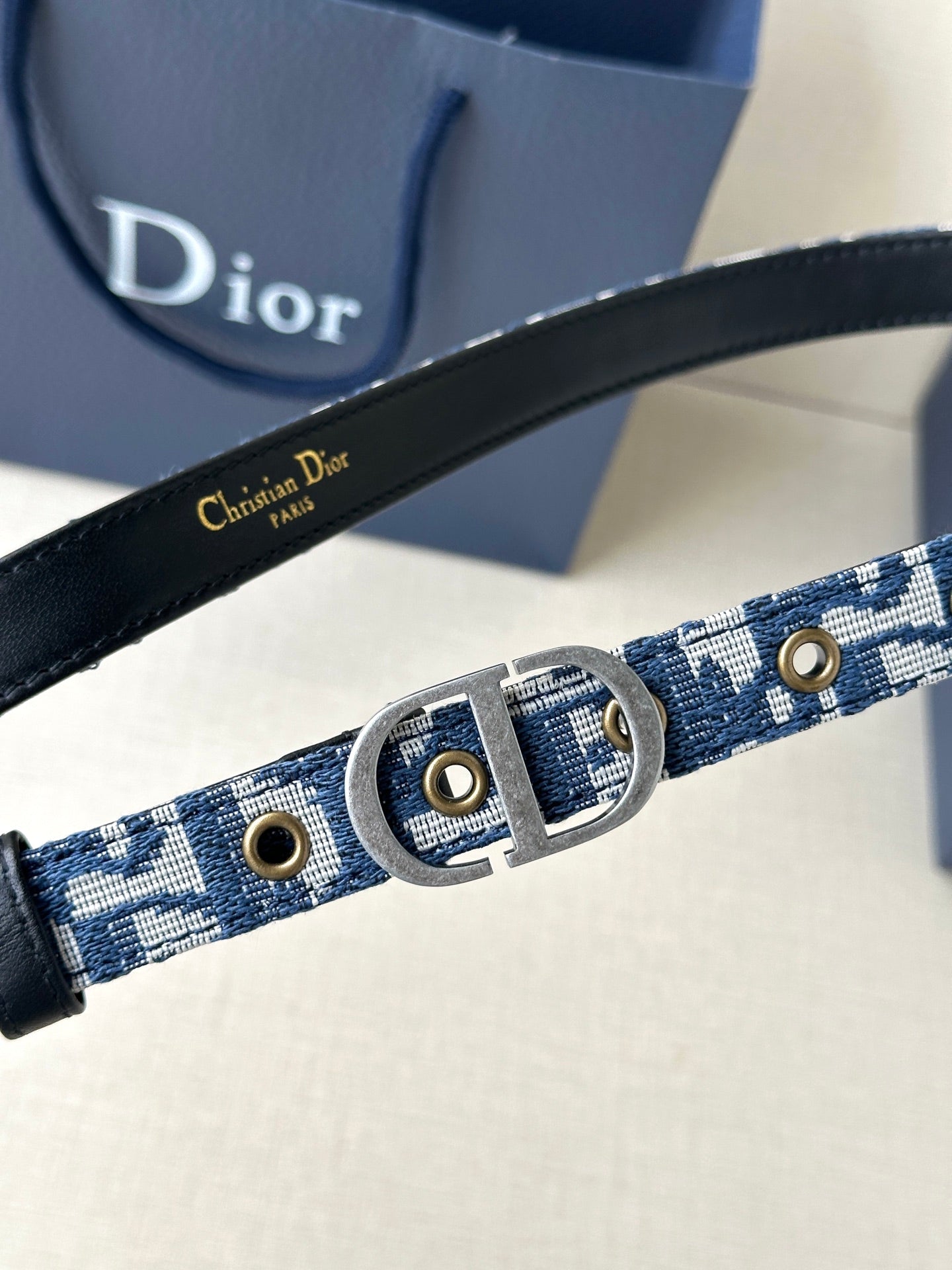 Dior belt close-up with blue monogram webbing, gold studs, and silver buckle.