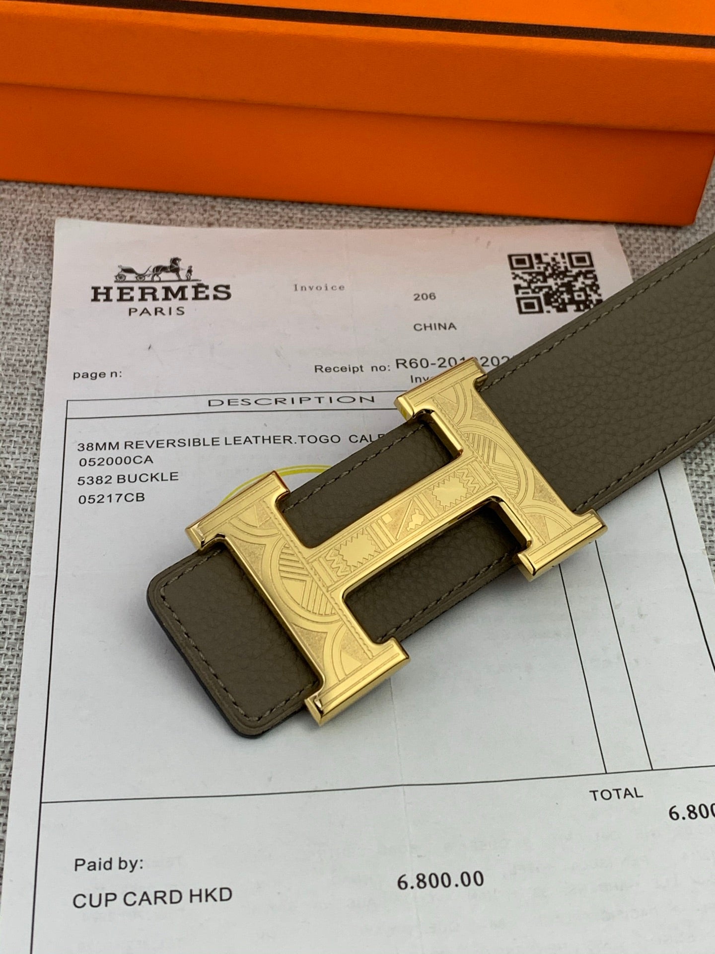 Leather belt close-up showing gold-tone H buckle on olive strap beside receipt.