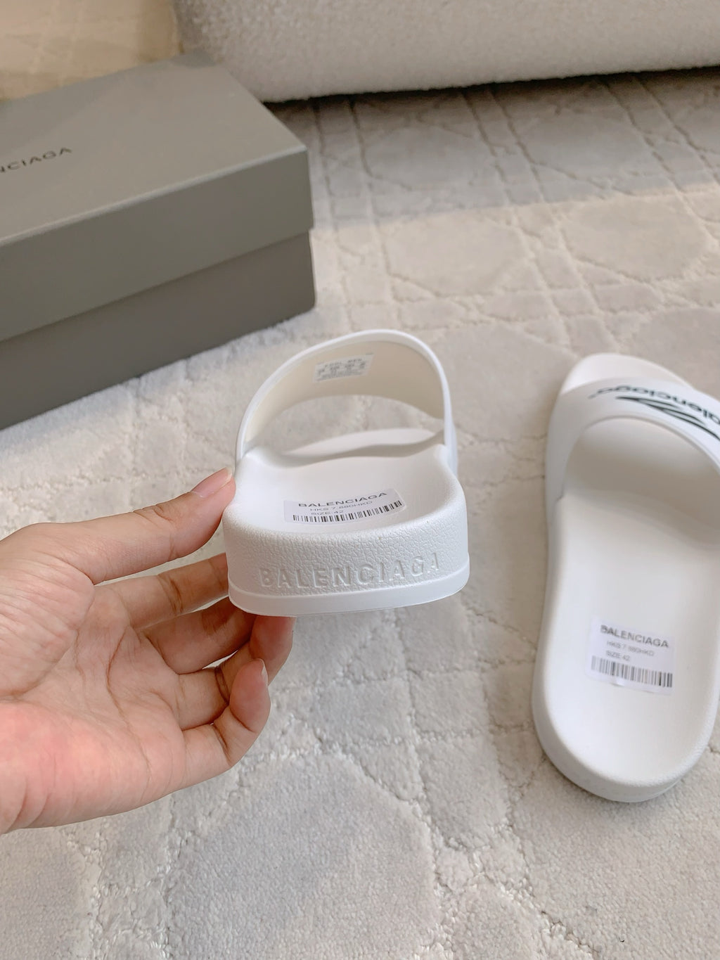 White Balenciaga slide sandals held in hand, with “balenciaga” strap text