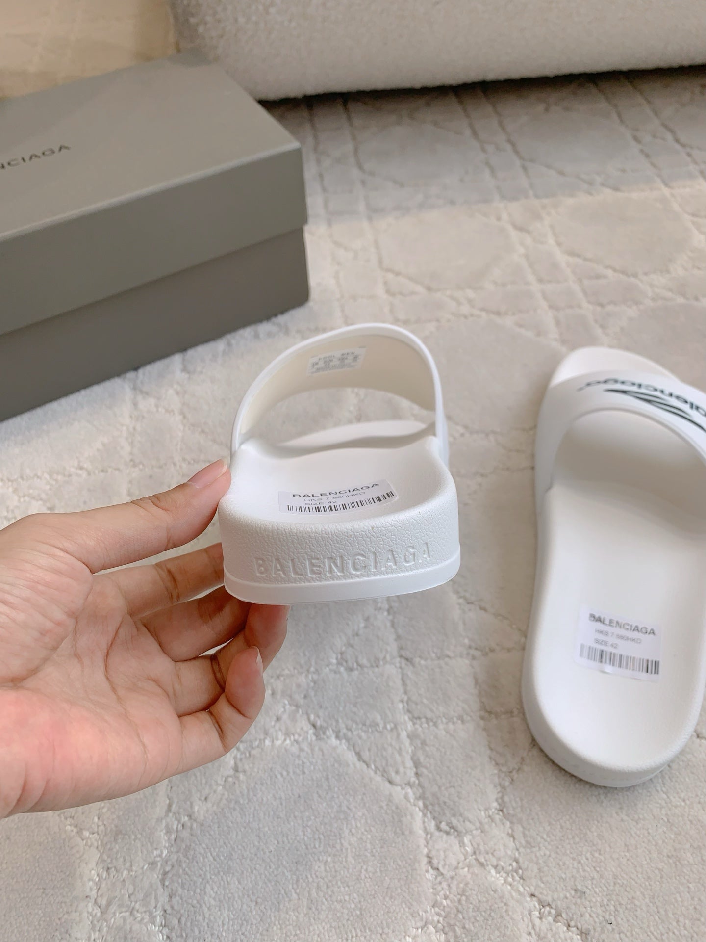 White Balenciaga slide sandals held in hand, with “balenciaga” strap text