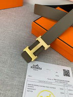 Leather belt close-up: light olive strap with gold-tone H buckle on orange gift box.