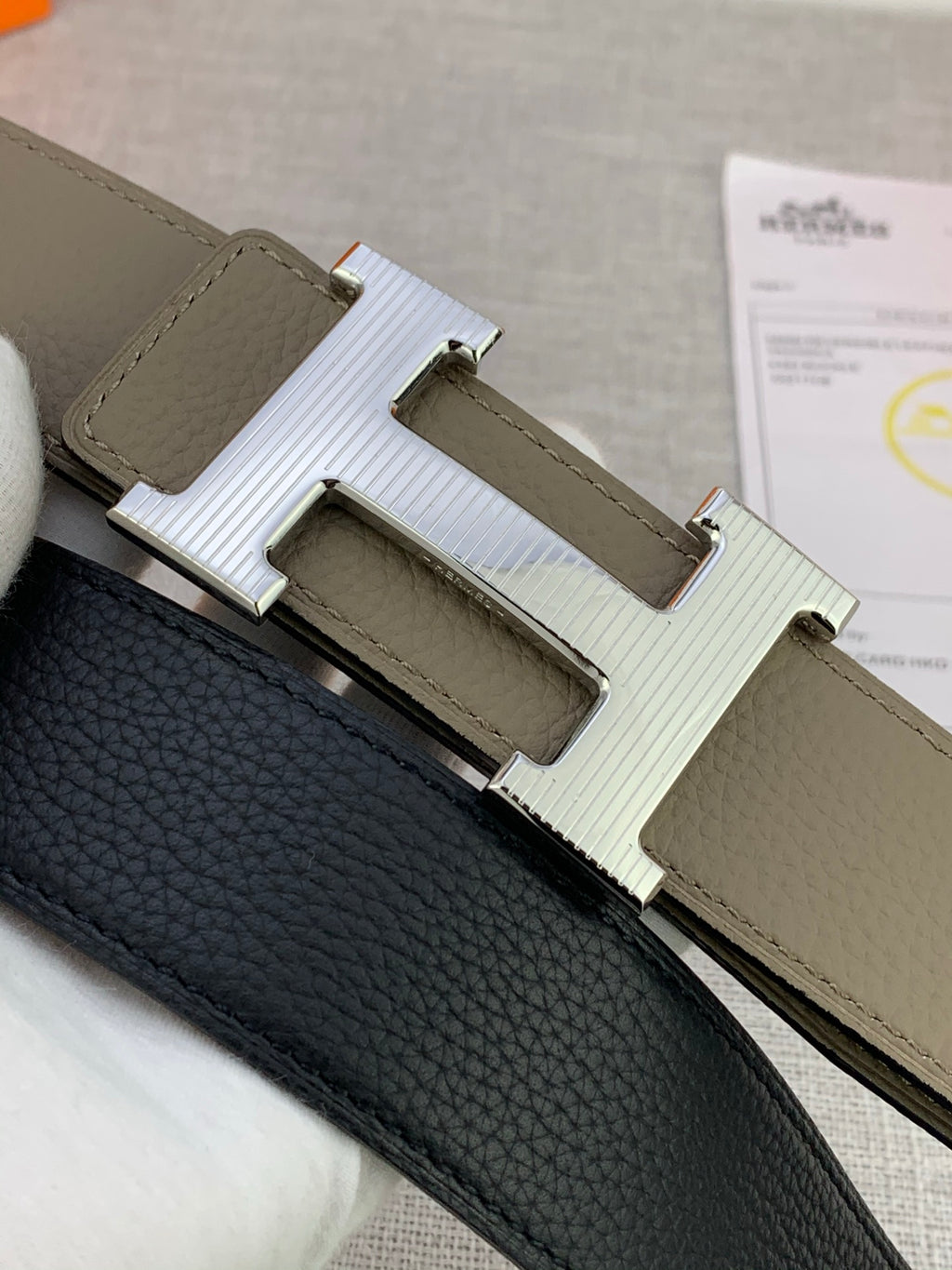 Close-up of reversible leather belt: black textured side with light olive strap and H buckle.