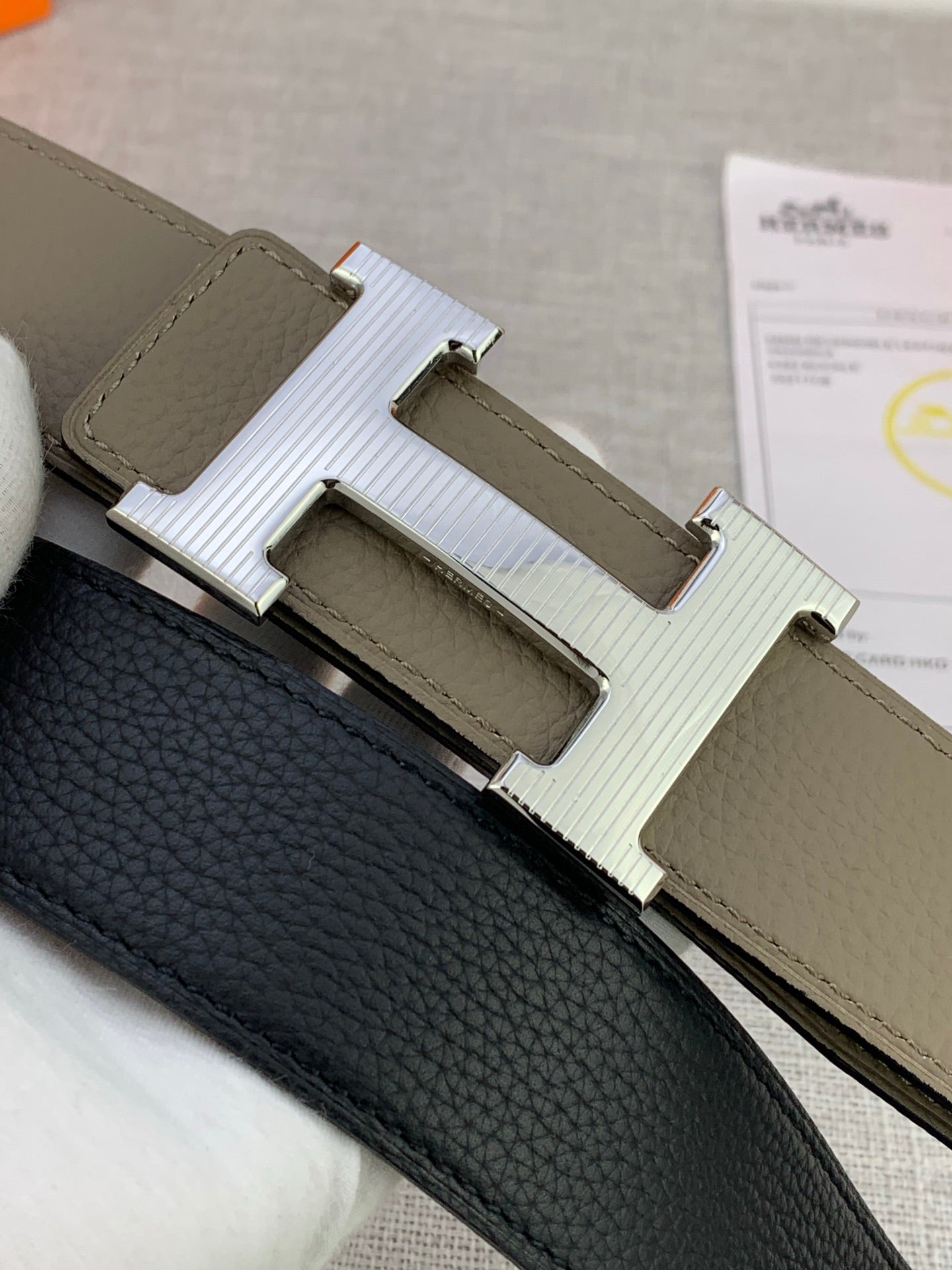 Close-up of reversible leather belt: black textured side with light olive strap and H buckle.