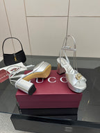 Silver metallic heeled sandals with gold logo hardware, open toe, ankle strap shown