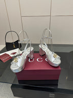 Silver open-toe platform sandals with gold logo hardware and tall slim heels, on display