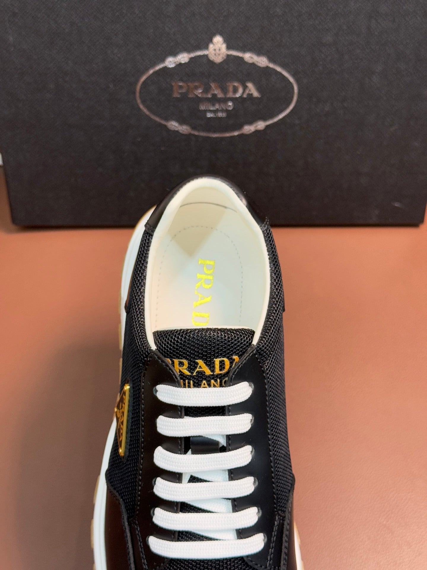 Prada unisex sneaker close-up, white laces and logo insole, dark upper.
