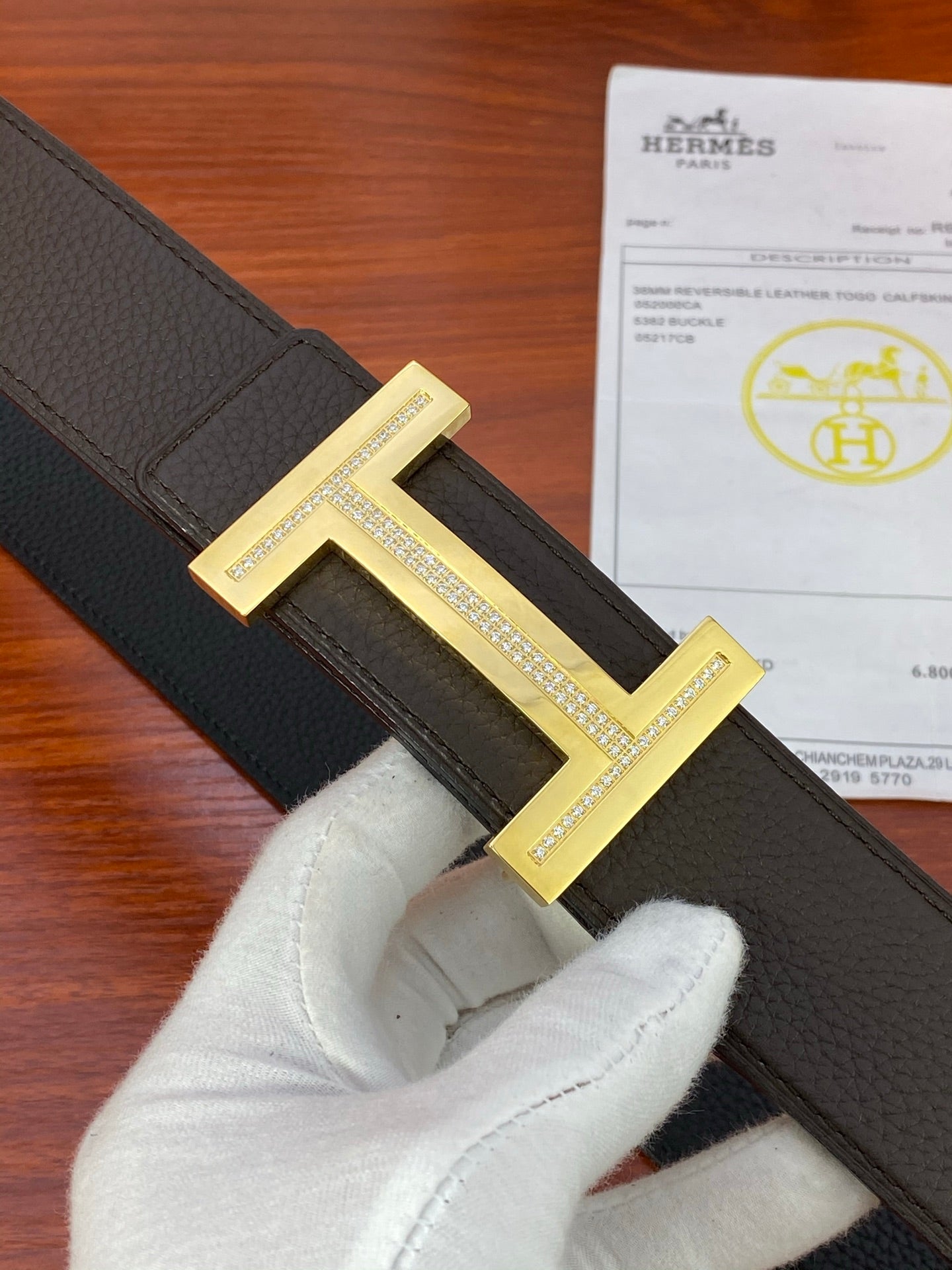 Black textured belt close-up with gold-tone H buckle and white-gloved hand