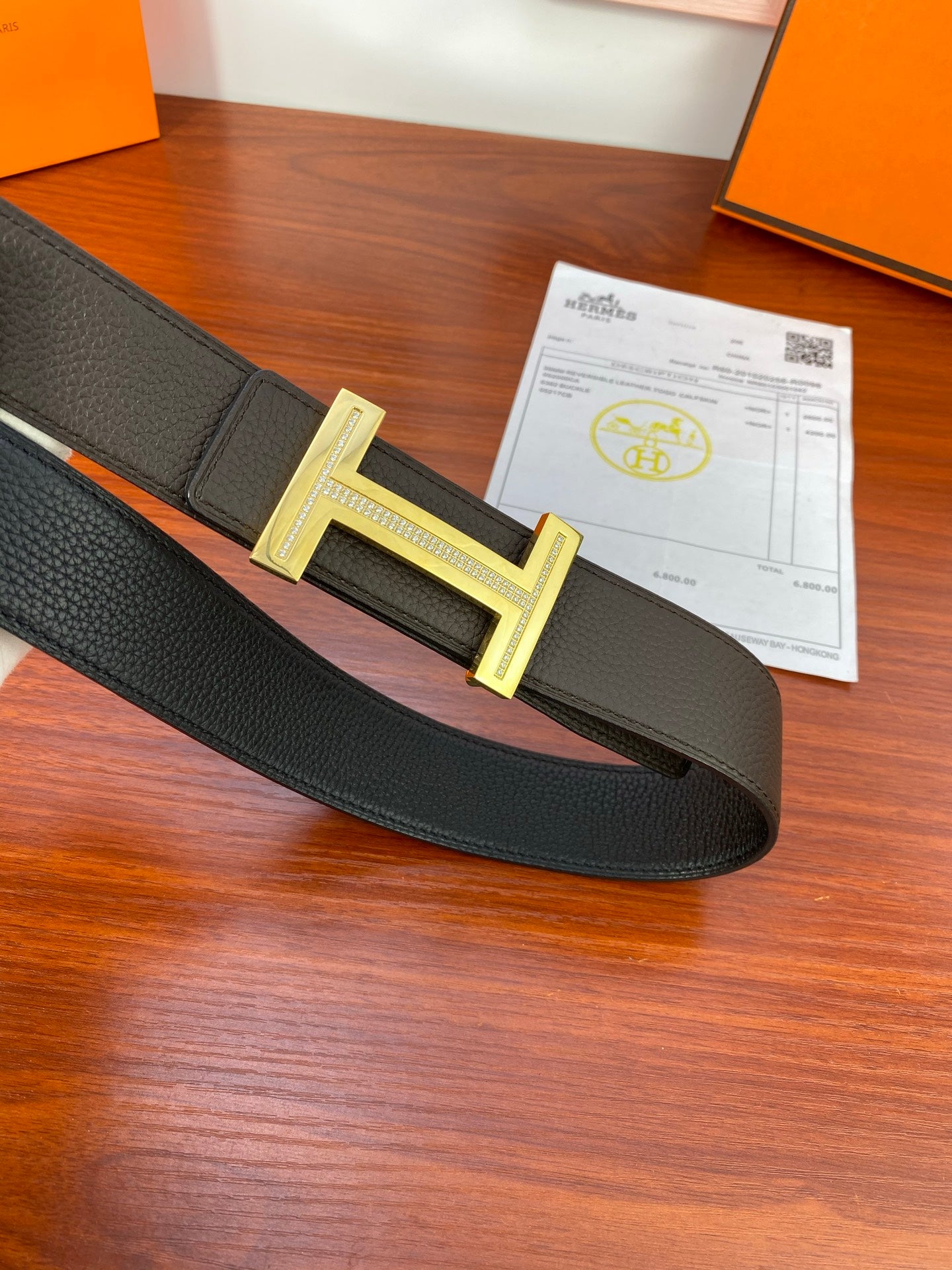 Black textured belt with gold-tone H buckle, laid flat with paperwork nearby