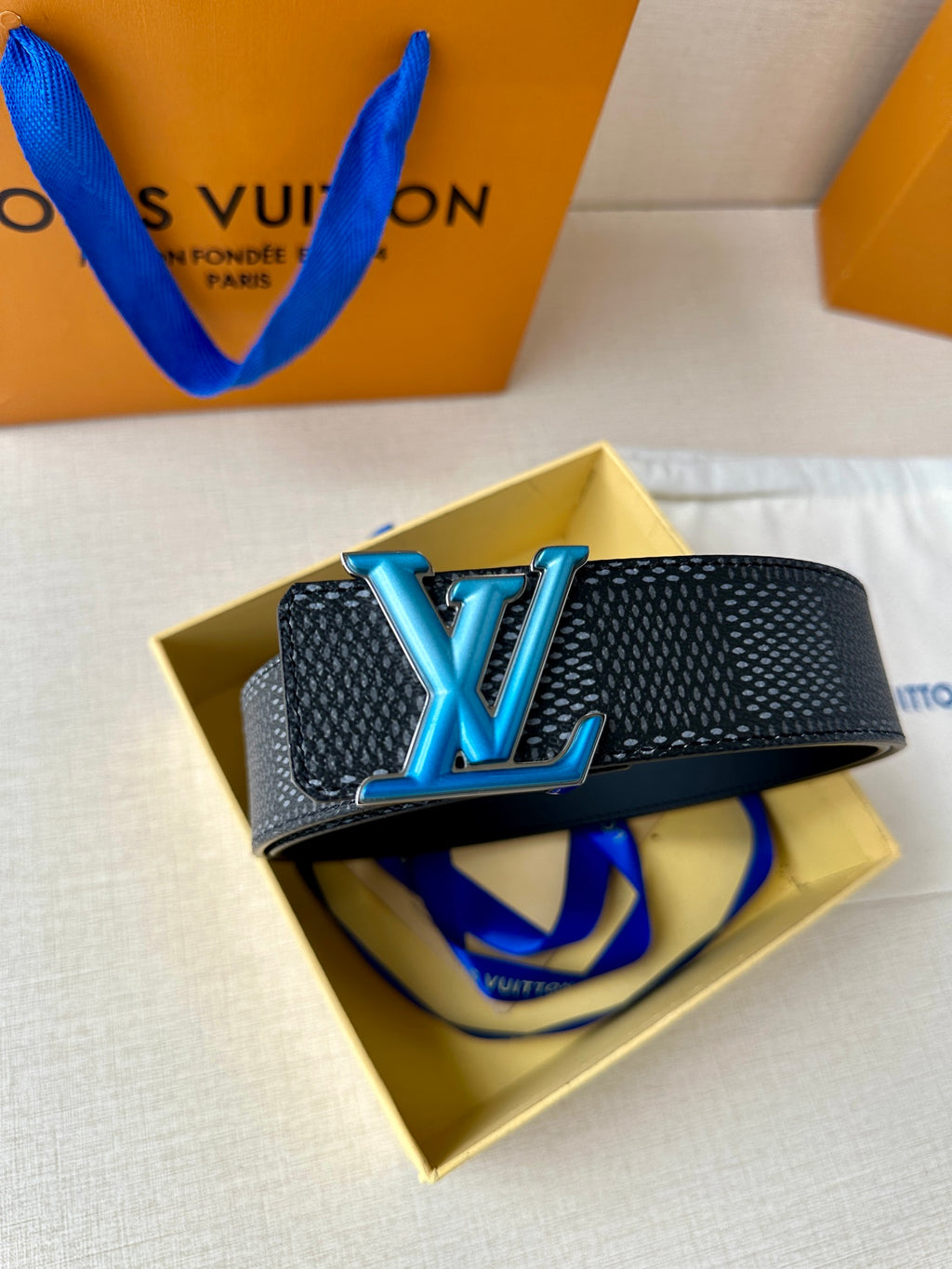 Louis Vuitton belt with dark textured strap and oversized blue LV buckle on yellow box.