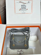 Gray leather handbag with gold-tone flap and hardware, shown in a gift box.