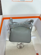 Gray leather handbag with top handle and flap front, side view in a box.