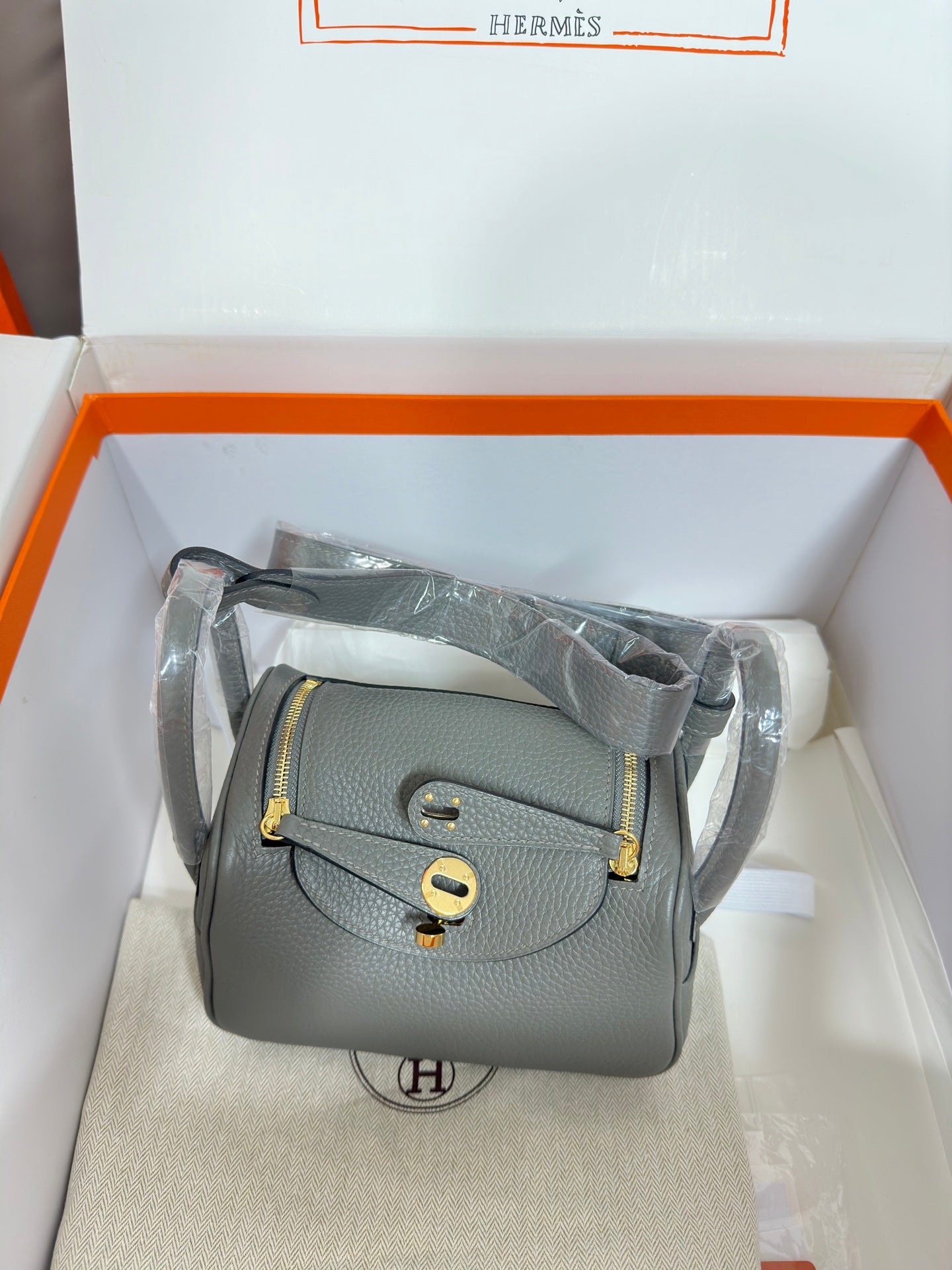 Gray leather crossbody handbag with gold-tone flap and shoulder strap, boxed.